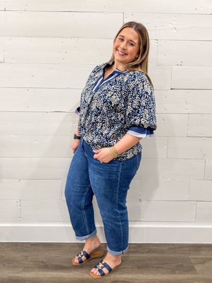 Woman wearing a blue floral blouse and jeans standing against a white brick wall.