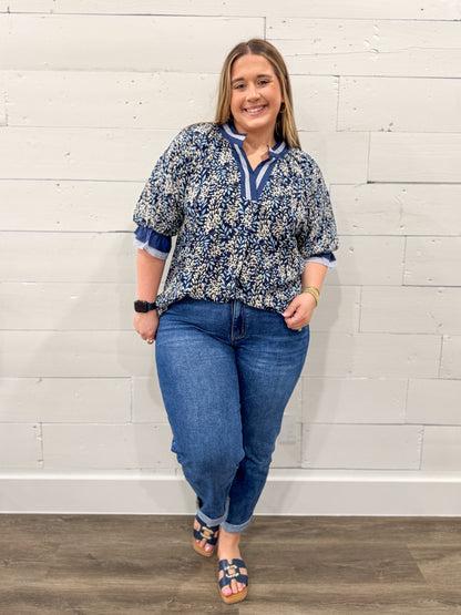 Woman wearing a blue floral blouse and jeans against a white brick wall.