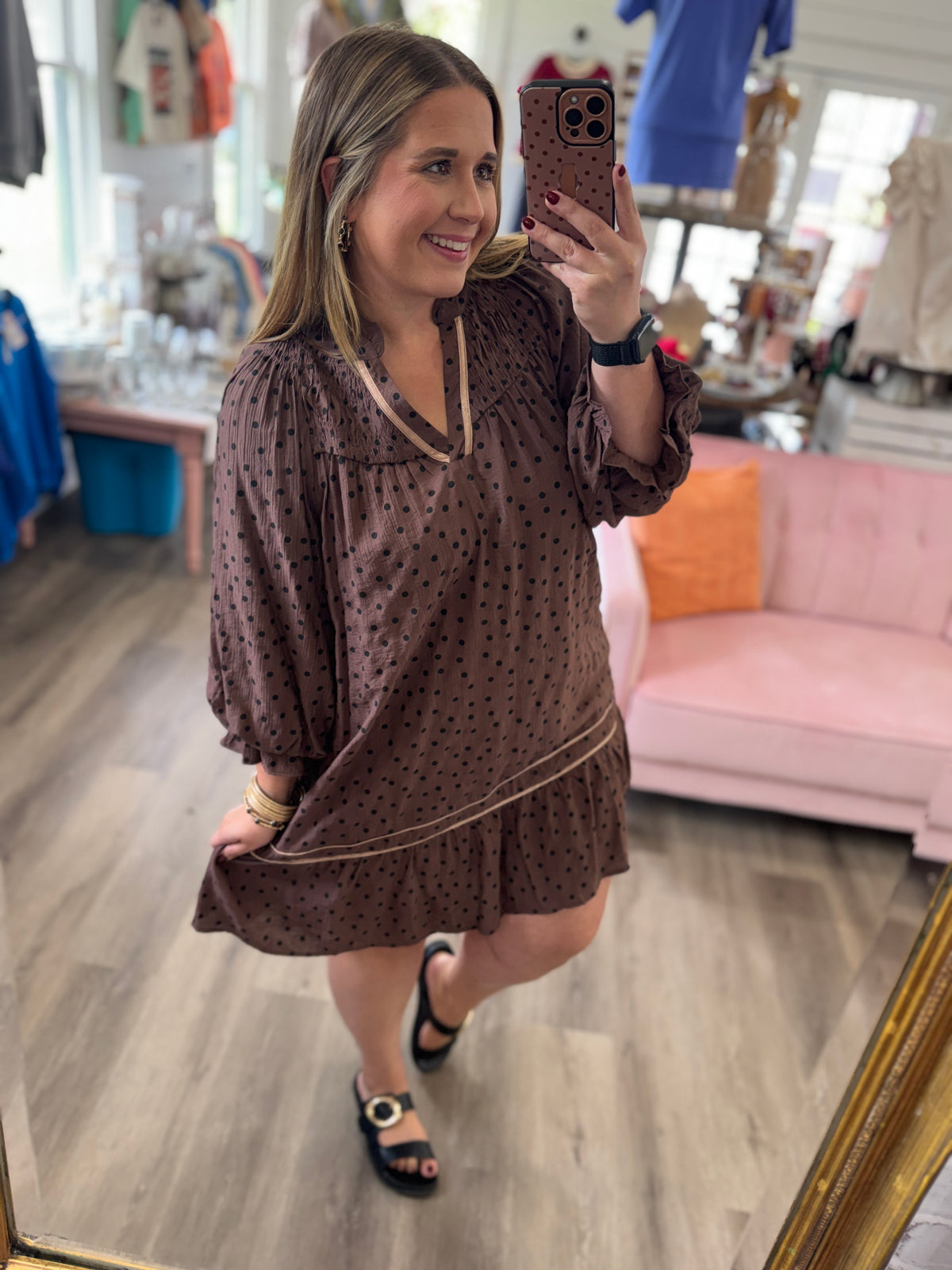 A woman takes a mirror selfie in a boutique wearing a brown long-sleeve tunic dress with black polka dots and beige trim. The background shows a pink sofa, wooden floors, and clothing racks.