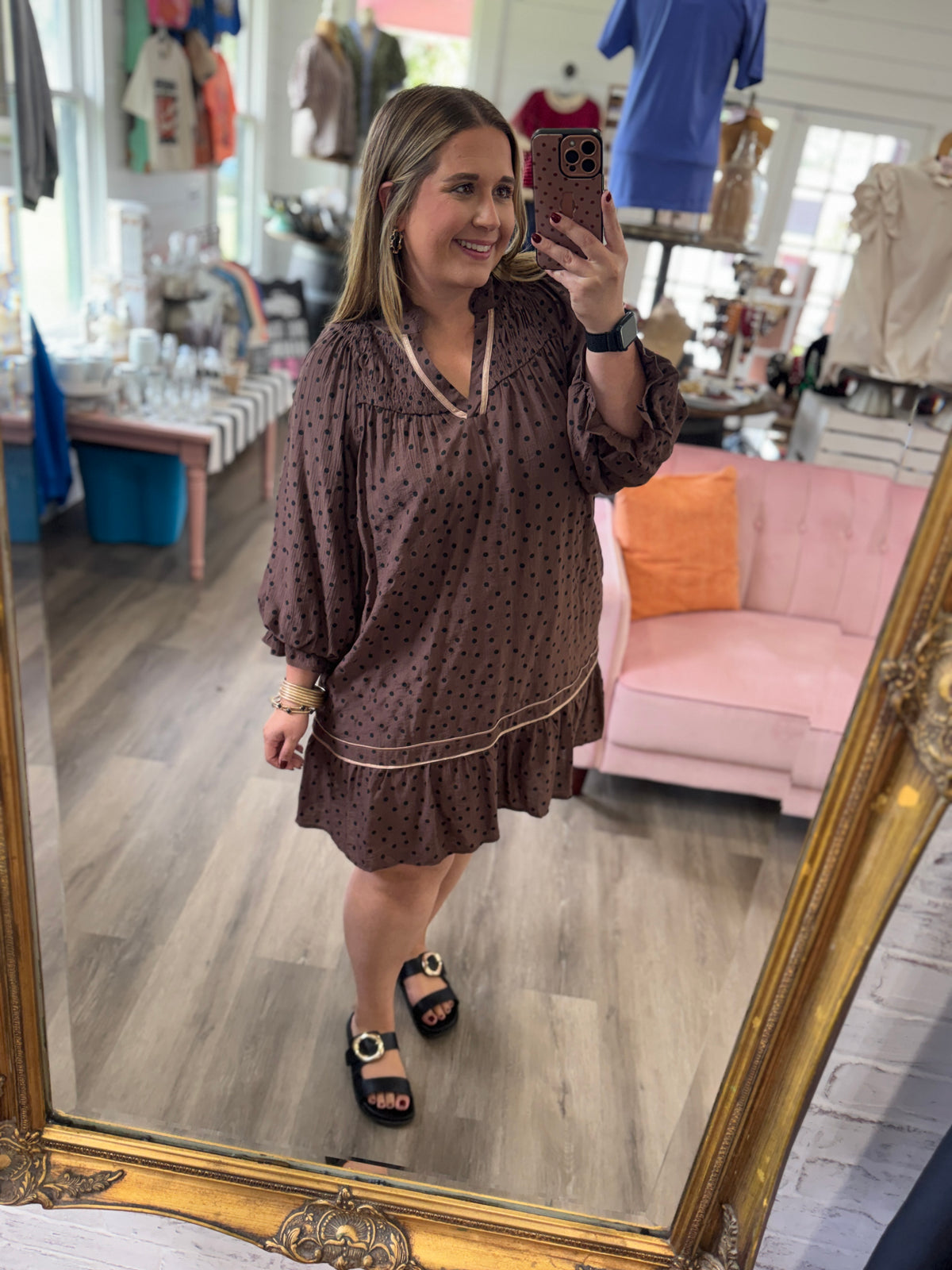 A woman takes a mirror selfie in a boutique shop, wearing a brown short-sleeve dress with small black polka dots and gold trim. The shop features a pink sofa, wooden floors, and clothing racks in the background.