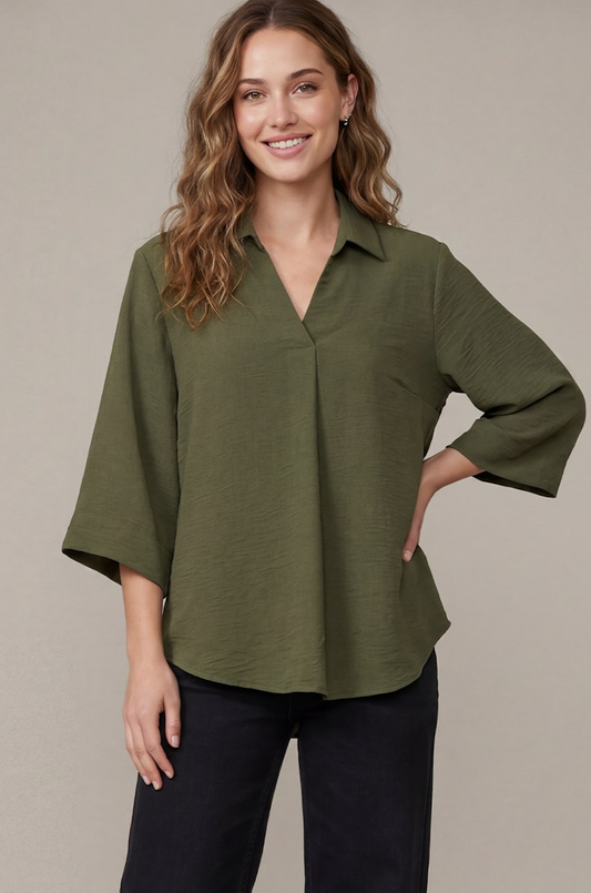 A woman wearing an olive green button-down blouse with three-quarter sleeves and dark pants, standing against a plain beige studio background.