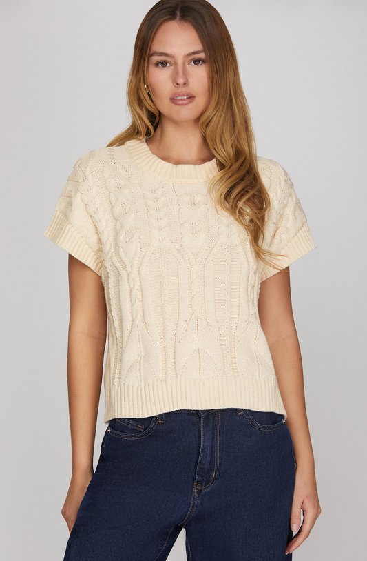 A woman wearing a cream-colored short-sleeve cable-knit sweater and dark blue jeans, standing against a light gray studio background.