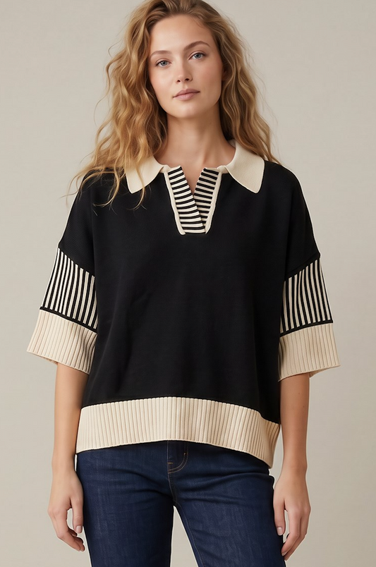 A woman modeling a black knit top with cream ribbed collar, sleeve cuffs, and hem, featuring black and cream vertical stripes on the chest and sleeves, paired with dark blue jeans against a neutral beige background.