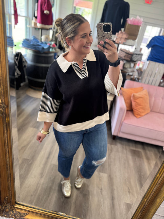 A woman takes a mirror selfie in a boutique wearing a black knit sweater with cream ribbed collar, hem, and sleeve accents, paired with blue jeans and silver sneakers.