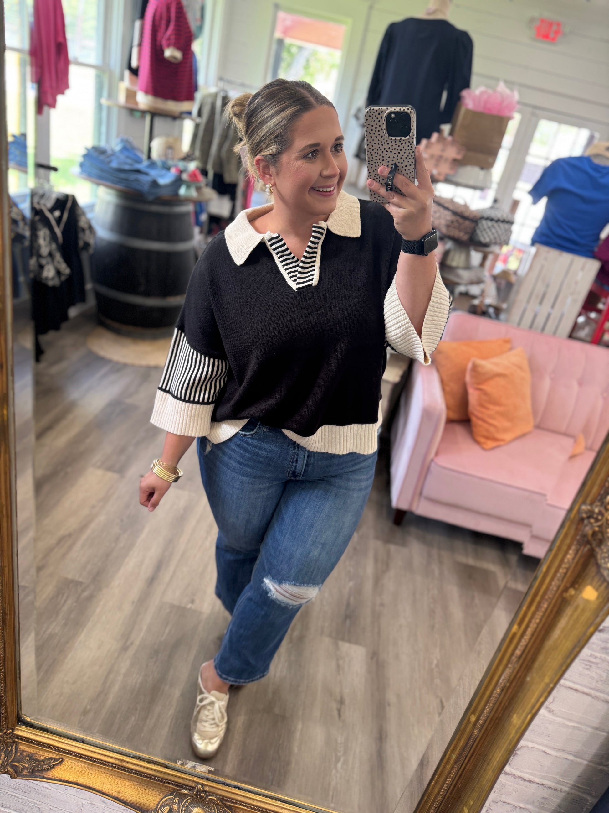 A woman takes a mirror selfie in a boutique while wearing a black knit sweater with cream and white striped details on the collar and sleeves, paired with blue jeans and gold sneakers.