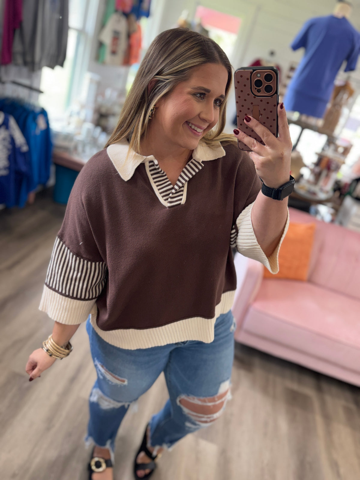 A woman takes a mirror selfie in a clothing boutique, wearing a brown and cream knit polo sweater with striped details on the collar and sleeves, paired with distressed blue jeans.