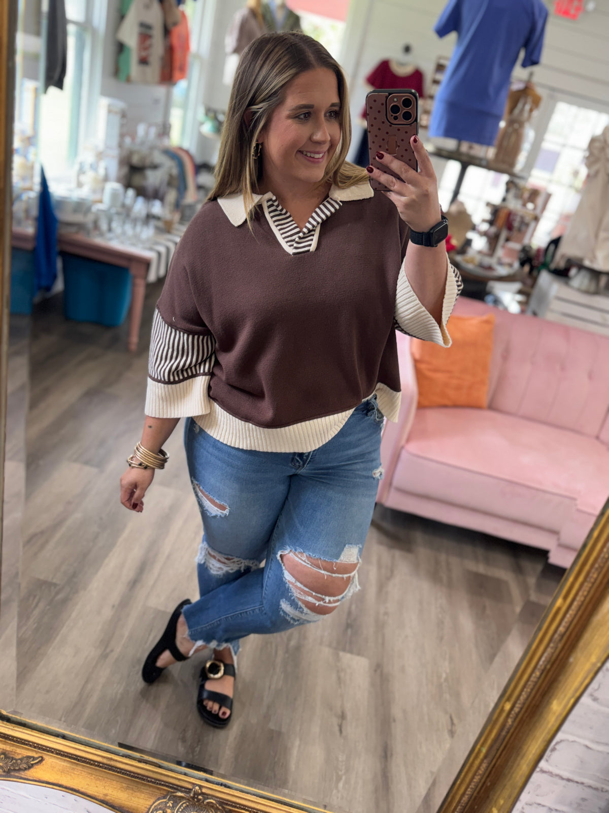 A woman takes a mirror selfie in a boutique shop, wearing a brown and cream knit sweater vest with striped sleeves, blue ripped jeans, and black sandals.