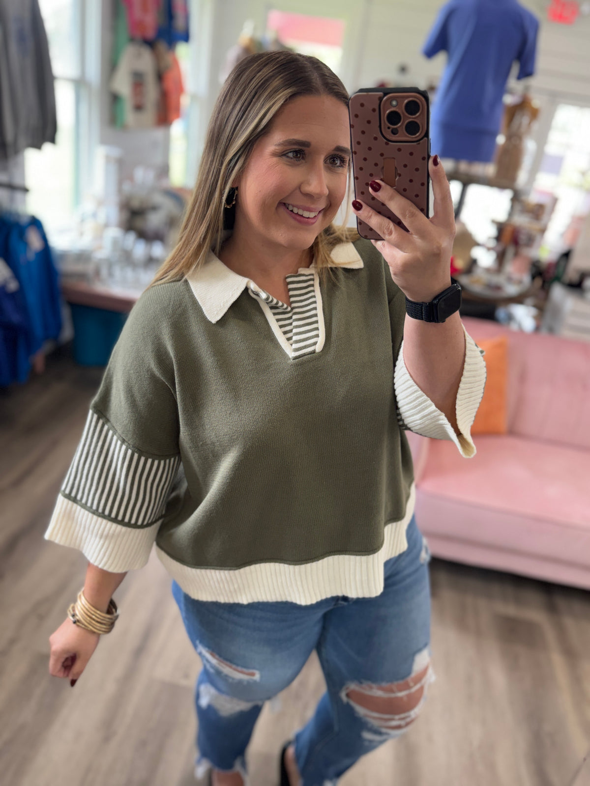 A woman takes a mirror selfie in a boutique while wearing a color-blocked knit sweater with an olive green body, cream collar and hem, and striped three-quarter sleeves, paired with distressed blue jeans.
