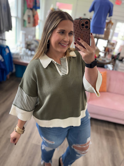 A woman takes a mirror selfie in a boutique while wearing a color-blocked knit sweater with an olive green body, cream collar and hem, and striped three-quarter sleeves, paired with distressed blue jeans.