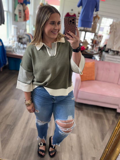 A woman takes a mirror selfie in a boutique wearing an olive green knit sweater with cream and striped accents, paired with distressed blue jeans and black sandals.