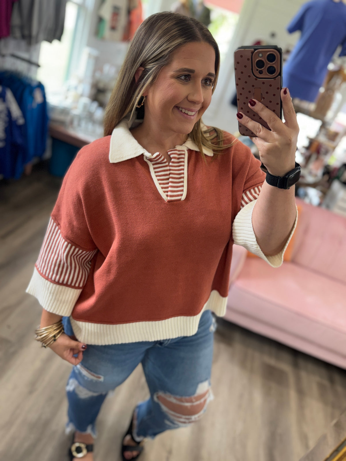 A woman takes a mirror selfie in a clothing store wearing a terracotta-colored knit polo sweater with cream and striped details, paired with distressed blue jeans.