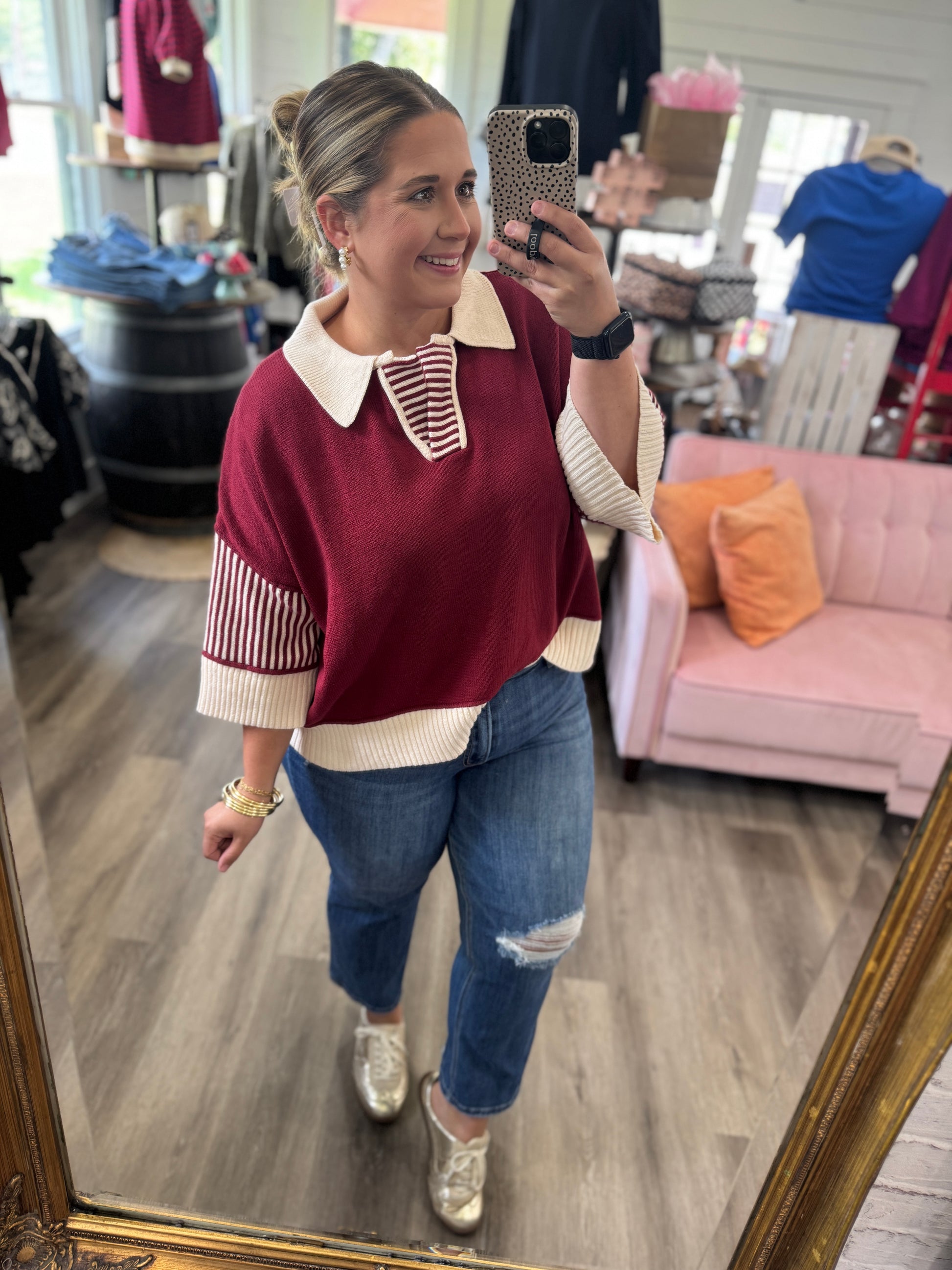 A woman takes a mirror selfie in a clothing boutique wearing a burgundy and cream knit sweater with striped details, blue ripped jeans, and gold sneakers.