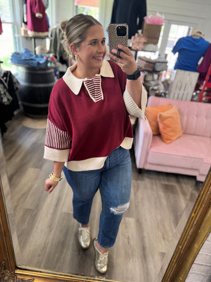 A woman takes a mirror selfie in a clothing boutique wearing a burgundy and cream knit sweater with striped details, blue ripped jeans, and gold sneakers.