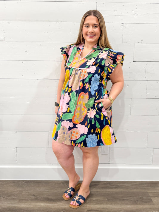 Woman wearing a colorful floral dress against a white brick wall.