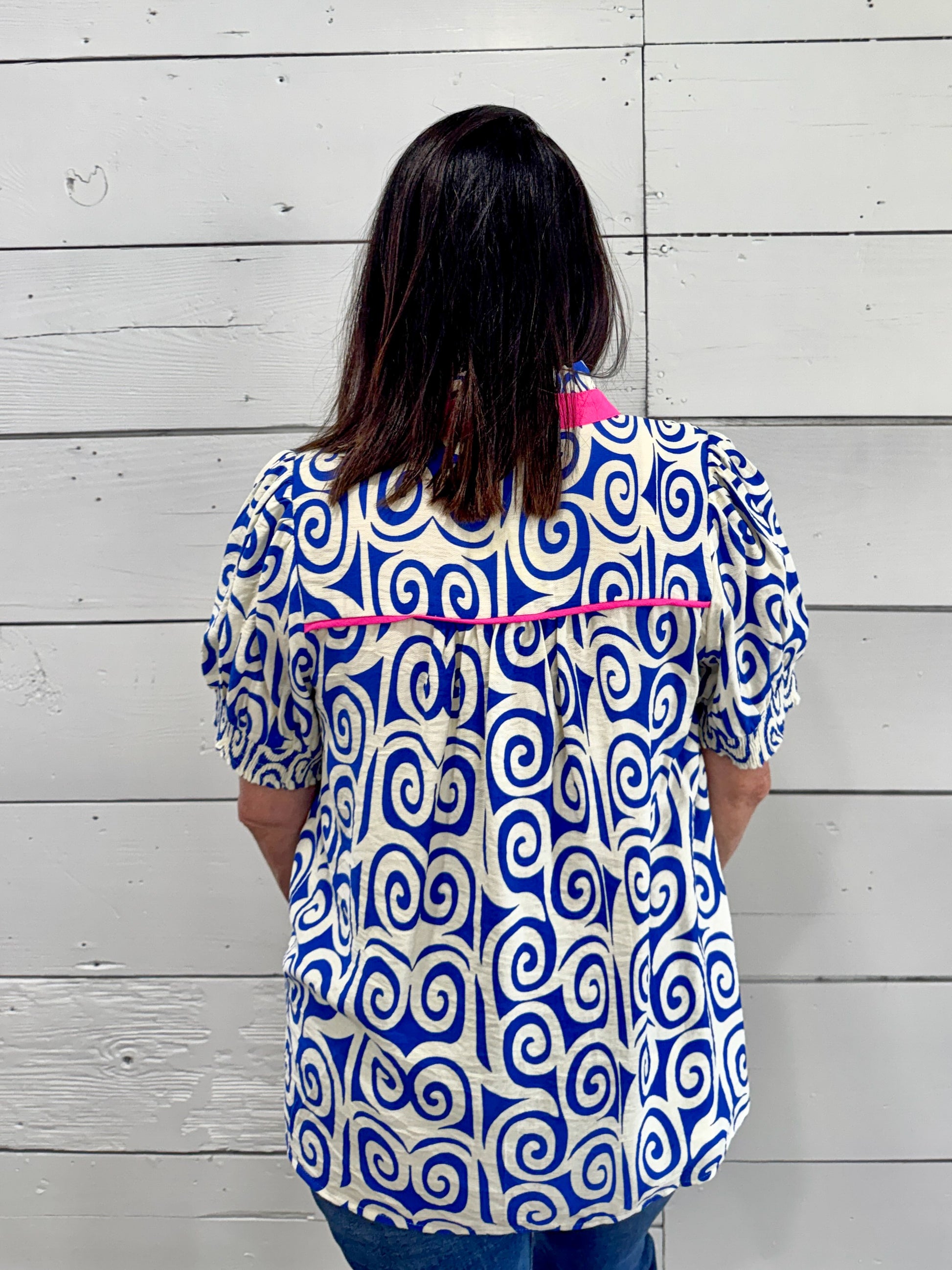A woman viewed from behind wearing a short-sleeve button-up blouse with a bold blue and white spiral pattern and bright pink trim, standing against a white wood plank wall.