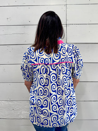 A woman viewed from behind wearing a short-sleeve button-up blouse with a bold blue and white spiral pattern and bright pink trim, standing against a white wood plank wall.