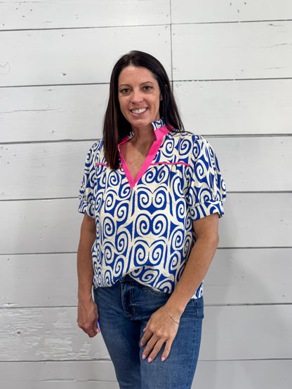 A woman wearing a blue and white patterned short-sleeve blouse with a bright pink inner collar, paired with blue jeans, standing against a white wood-paneled wall.