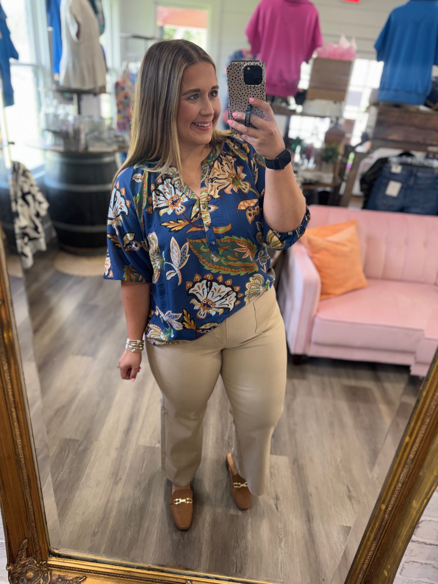 A woman takes a mirror selfie in a boutique, wearing a blue short-sleeve button-up blouse with a white, gold, and green floral pattern, paired with beige trousers and brown loafers.