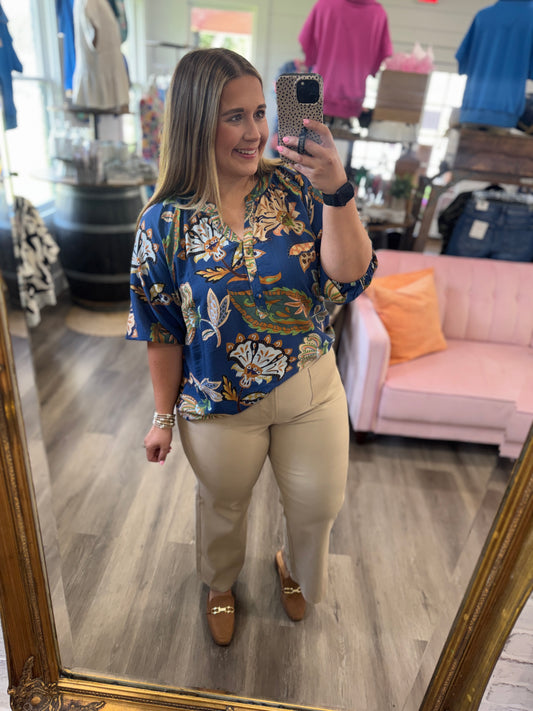A woman takes a mirror selfie in a boutique, wearing a blue short-sleeve button-up blouse with a white, gold, and green floral pattern, paired with beige trousers and brown loafers.