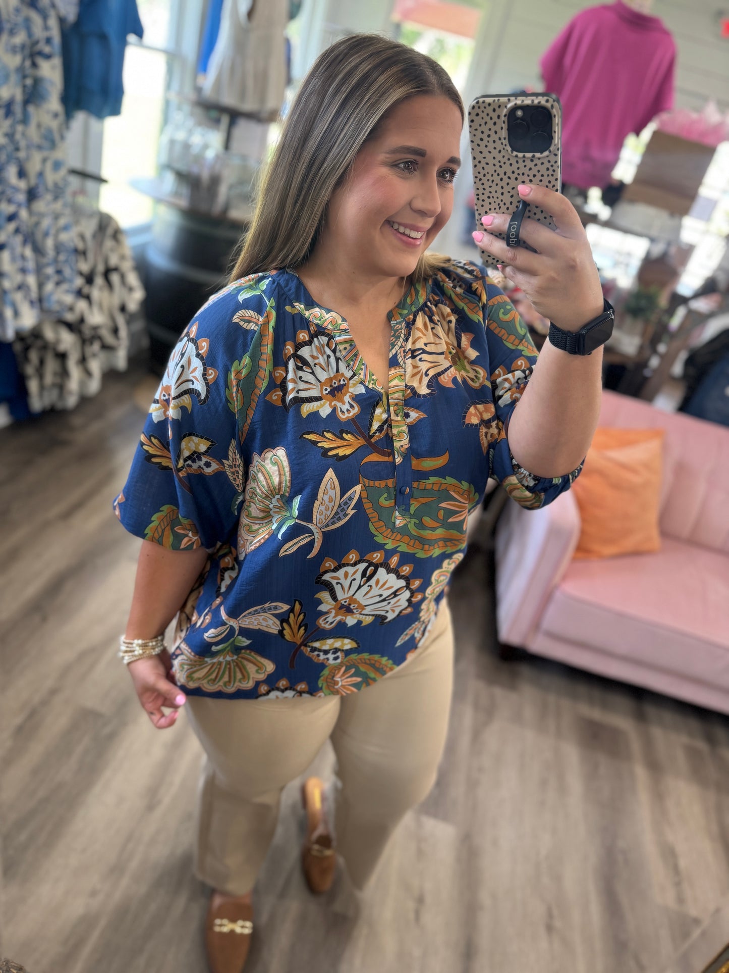A woman takes a mirror selfie in a boutique wearing a navy blue short-sleeve blouse with a beige, gold, and white floral pattern, paired with beige pants.