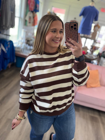 A woman taking a mirror selfie in a clothing store, wearing a brown and cream horizontally striped crewneck sweatshirt and blue jeans.