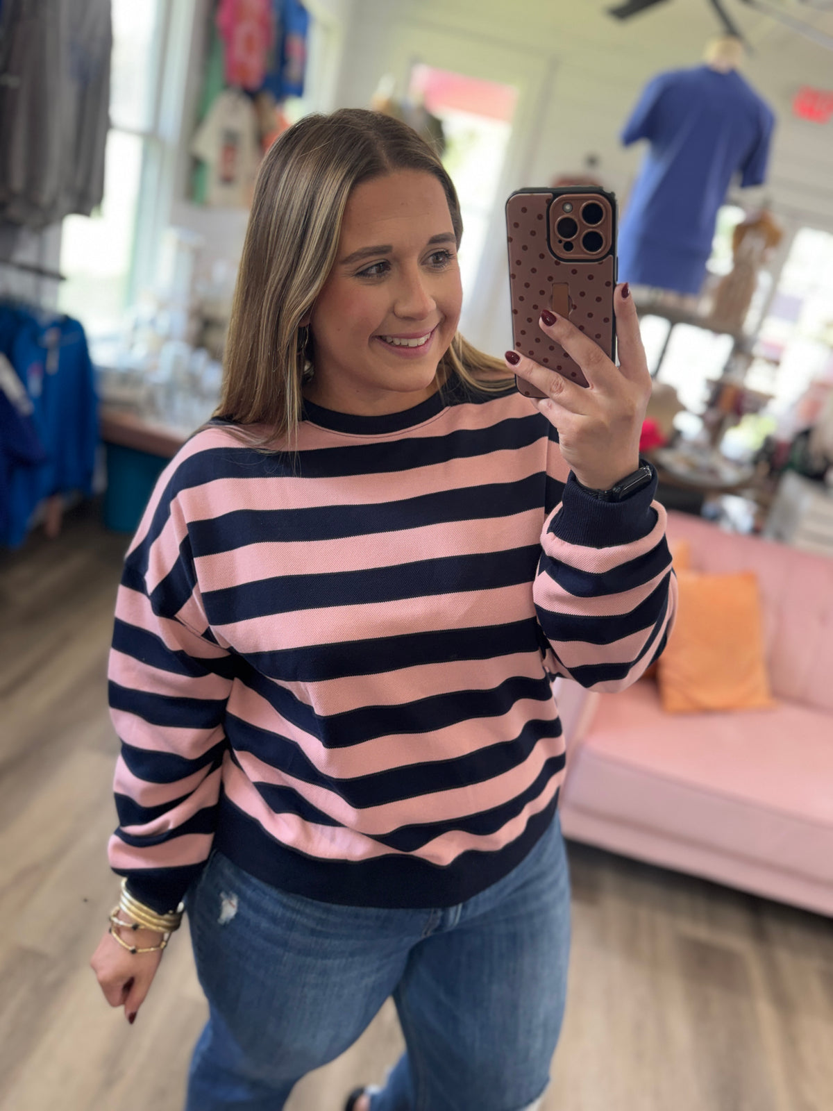 A woman takes a mirror selfie in a boutique while wearing a pink and navy blue horizontally striped crewneck sweatshirt and blue jeans.