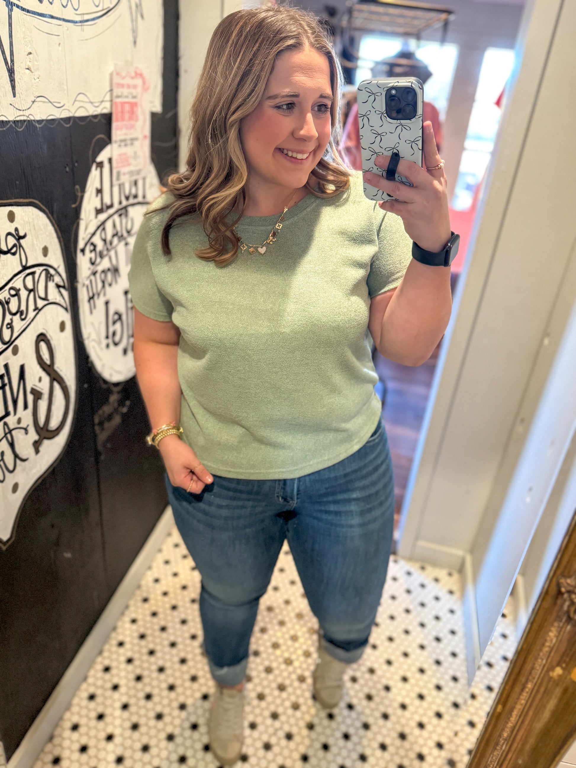 Woman taking a mirror selfie wearing a light green shirt and blue jeans.