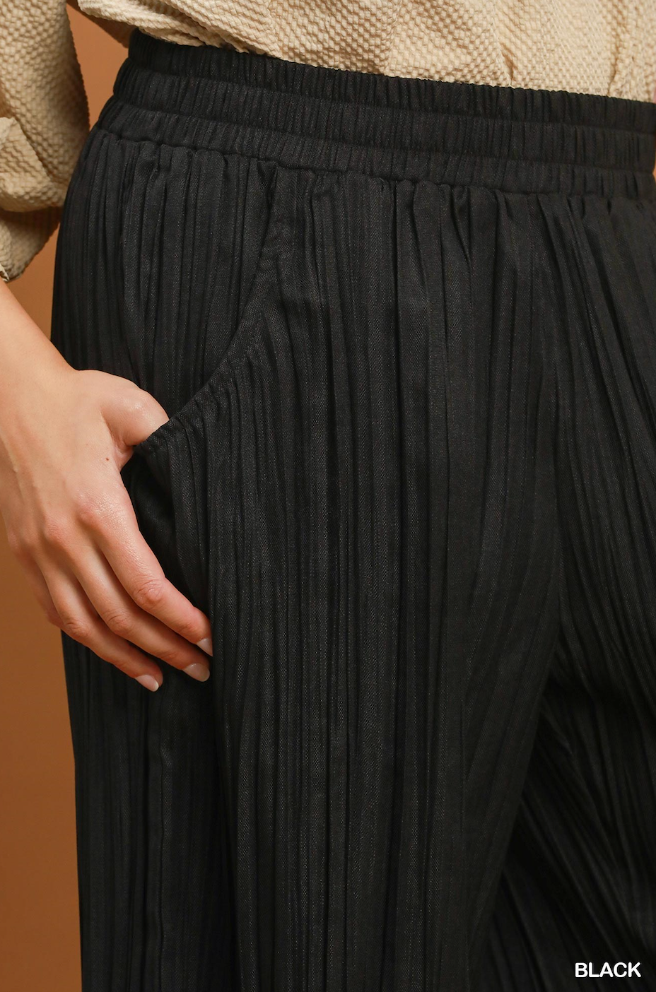 Close-up of a person wearing black pleated wide-leg trousers with a side pocket, paired with a beige knit top. A hand is visible holding the pocket edge.