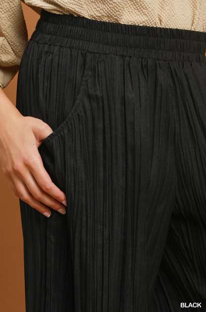 Close-up of a person wearing black pleated wide-leg trousers with a side pocket, paired with a beige knit top. A hand is visible holding the pocket edge.