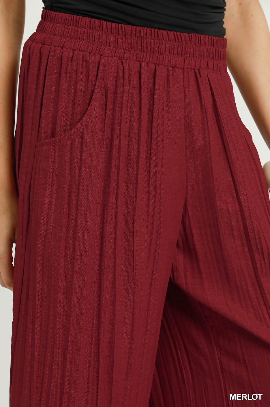 Close-up of a person wearing pleated wide-leg trousers in a deep merlot red fabric, paired with a black top. The image includes a 'MERLOT' text label in the bottom right corner.