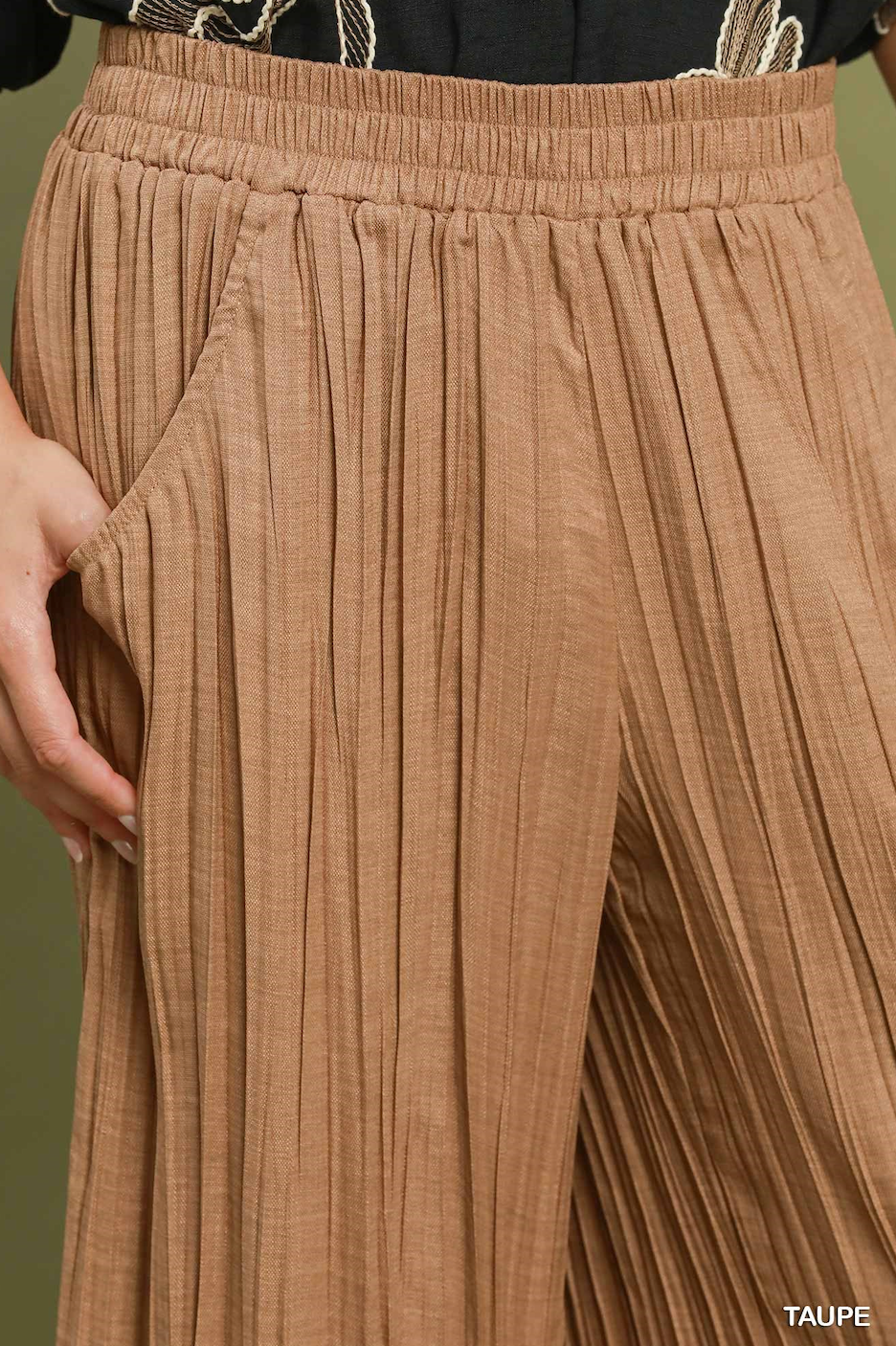 Close-up of pleated wide-leg pants in taupe, showing the elastic waistband, side pocket, and textured fabric. A hand is visible at the side, and the word 'TAUPE' appears in the bottom right corner.