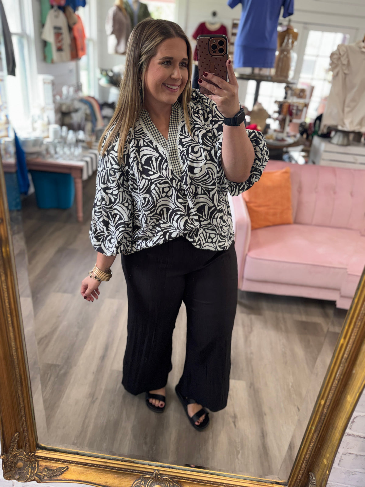 A woman takes a mirror selfie in a boutique, wearing a black and white abstract patterned wrap blouse with three-quarter sleeves and black wide-leg pants.