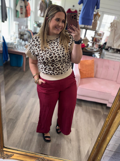 A woman takes a mirror selfie in a boutique, wearing a beige and black leopard print ruffle-sleeve crop top with a cream waistband and high-waisted burgundy wide-leg pleated pants.