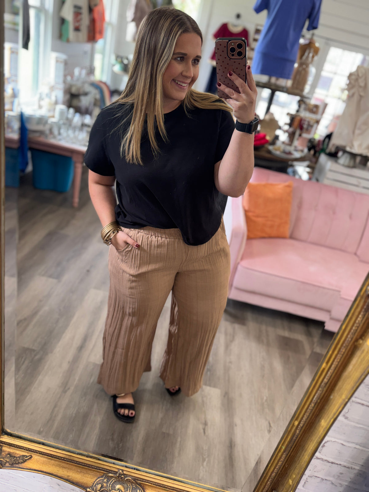 A woman takes a mirror selfie in a boutique, wearing a black short-sleeve t-shirt and pleated tan wide-leg pants. The background shows a pink sofa, clothing racks, and a gold-framed mirror.
