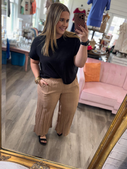 A woman takes a mirror selfie in a boutique, wearing a black short-sleeve t-shirt and pleated tan wide-leg pants. The background shows a pink sofa, clothing racks, and a gold-framed mirror.