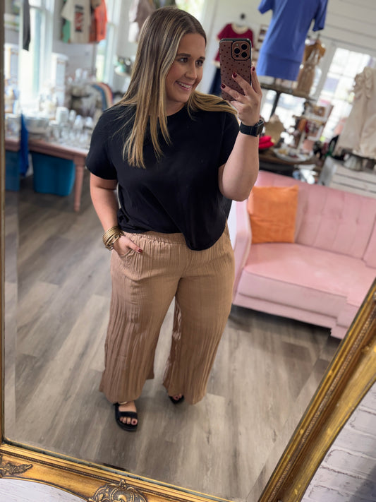 A woman takes a mirror selfie in a boutique, wearing a black short-sleeve t-shirt and pleated tan wide-leg pants. The background shows a pink sofa, clothing racks, and a gold-framed mirror.
