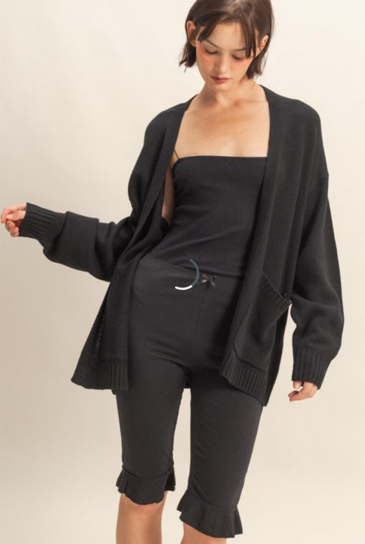 Woman wearing a black cardigan over a black top and capri pants on a beige background