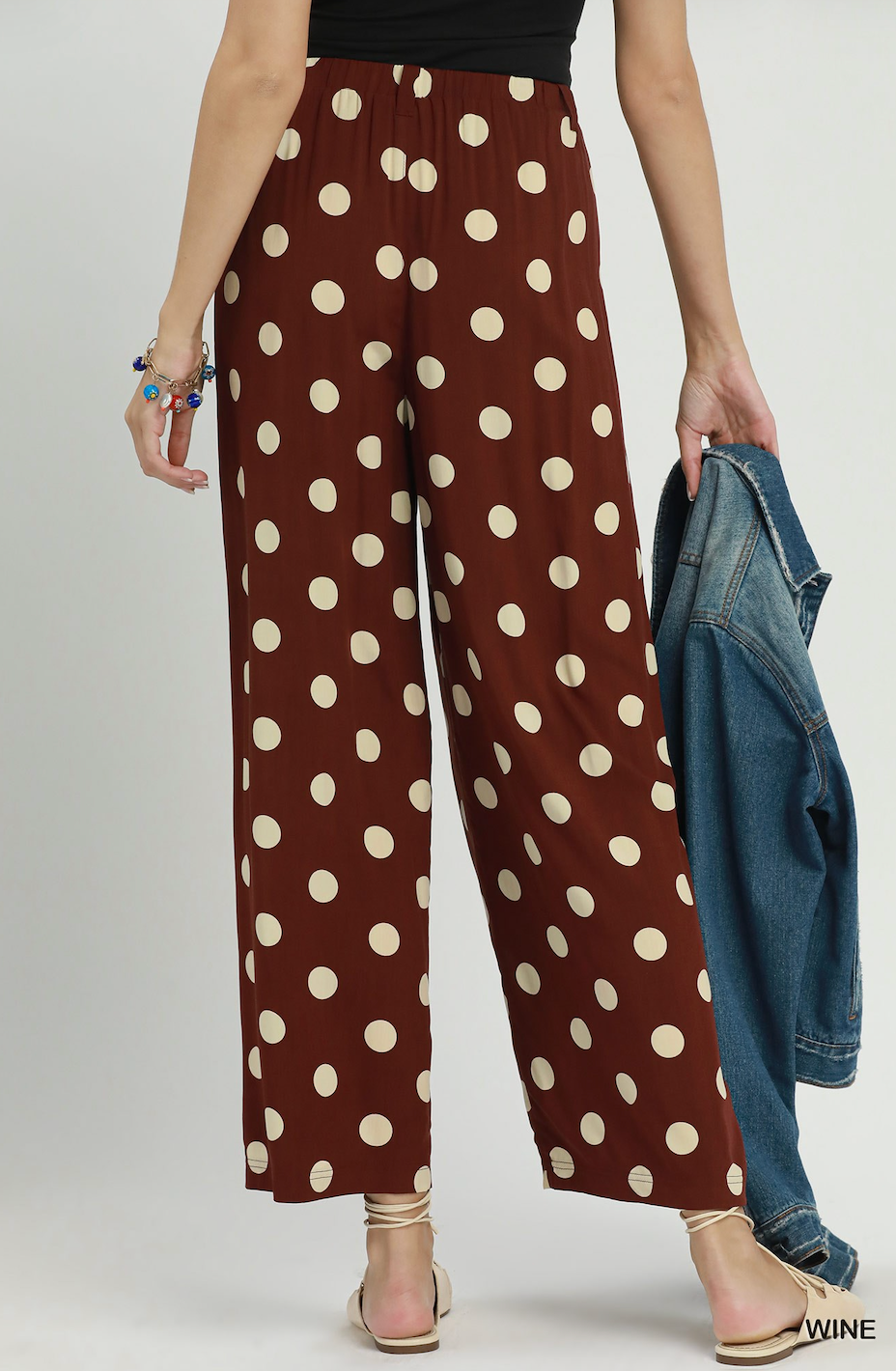 A model is shown from the waist down wearing brown wide-leg pants with a beige polka dot pattern. She is holding a blue denim jacket and wearing beige strappy sandals against a light gray studio background.