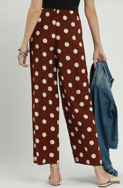 A model is shown from the waist down wearing brown wide-leg pants with a beige polka dot pattern. She is holding a blue denim jacket and wearing beige strappy sandals against a light gray studio background.