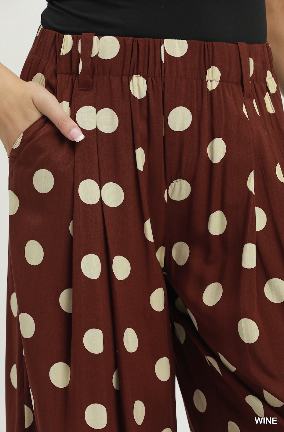 Close-up of a person wearing wide-leg pleated trousers in a wine brown color with large cream polka dots. A hand is visible in the pocket, and a small 'WINE' text label appears in the bottom right corner.
