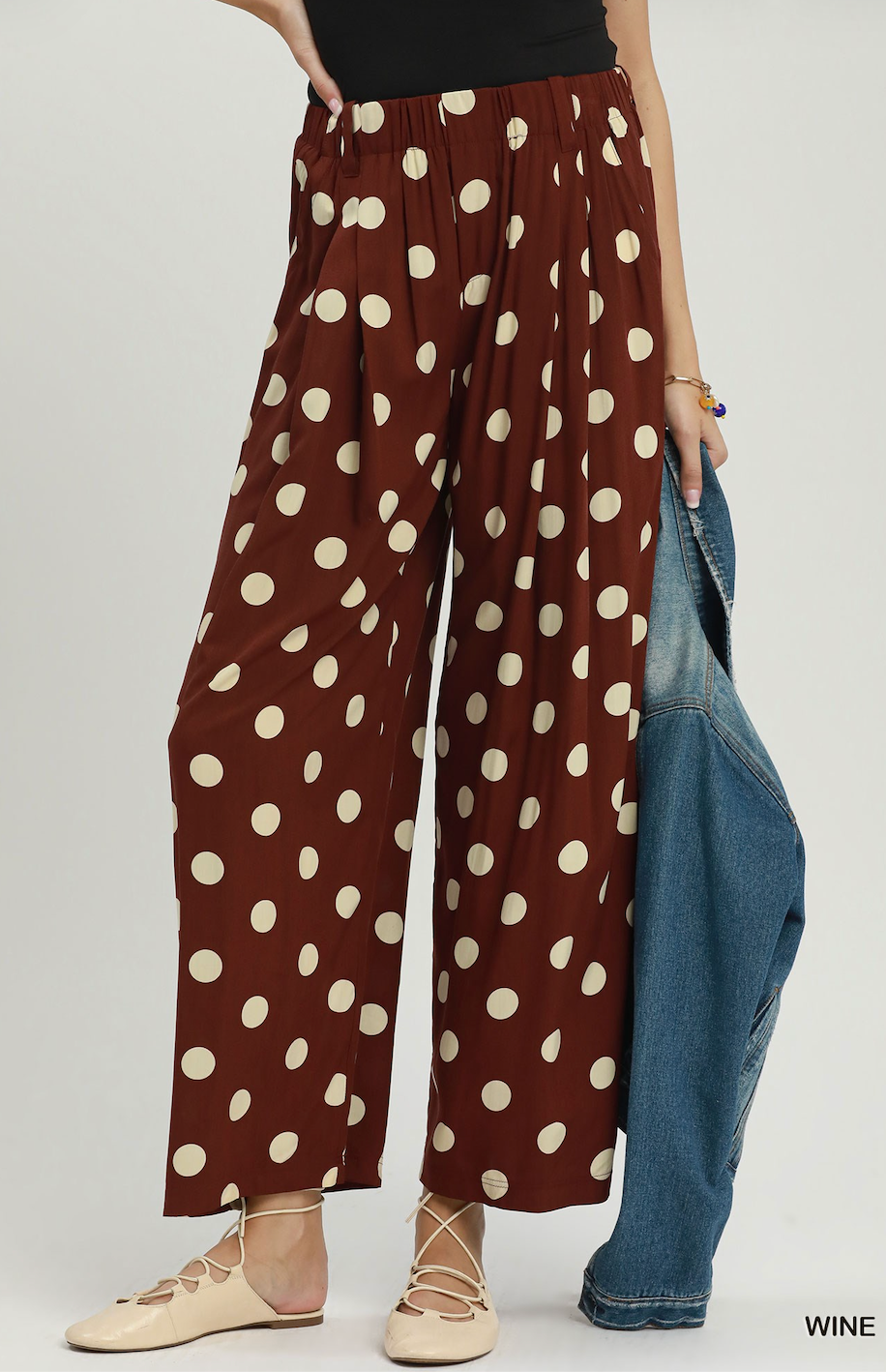 A model wears wide-leg brown pants with a cream polka dot pattern, holding a blue denim jacket. The background is a clean, light gray studio backdrop.