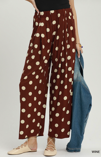 A model wears wide-leg brown pants with a cream polka dot pattern, holding a blue denim jacket. The background is a clean, light gray studio backdrop.