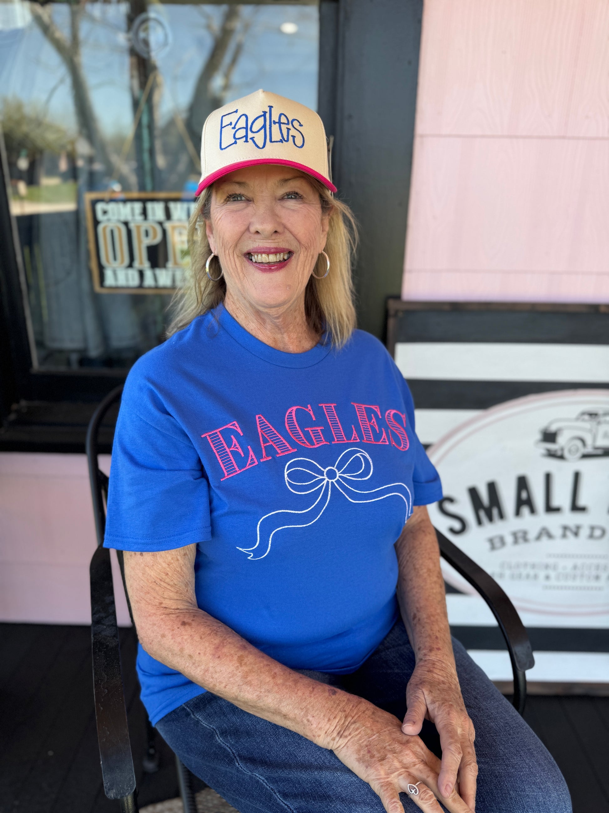 Woman wearing a blue 'EAGLES' t-shirt and beige cap sitting in front of a store entrance.