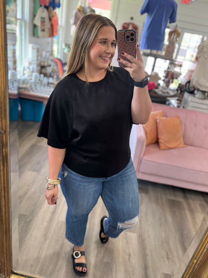 A woman takes a mirror selfie in a boutique while wearing a black short-sleeve top, blue ripped jeans, and black slide sandals. The shop has a pink sofa, wood floors, and clothing racks in the background.