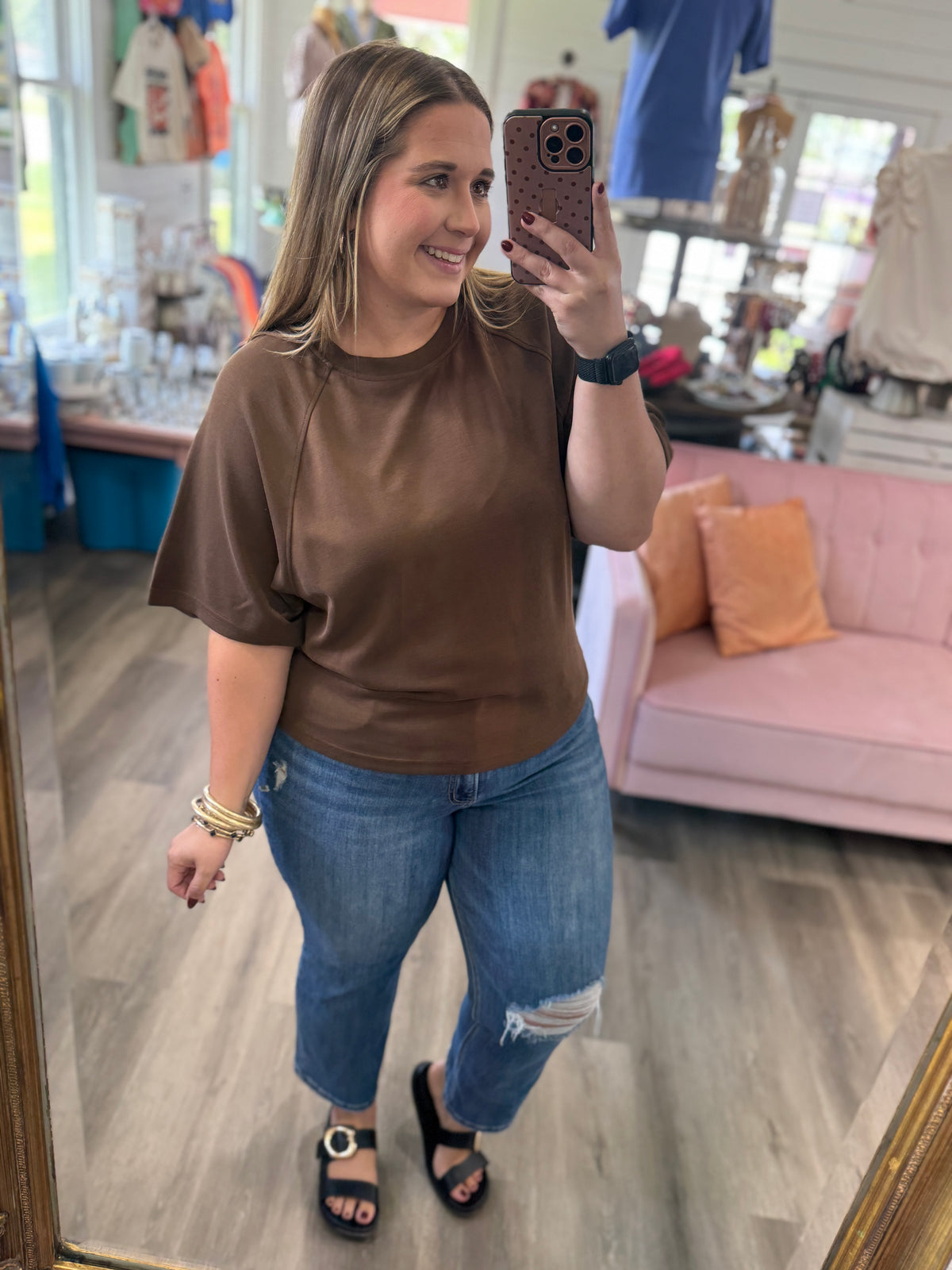 A woman takes a mirror selfie in a boutique, wearing a brown short-sleeve top and blue jeans.