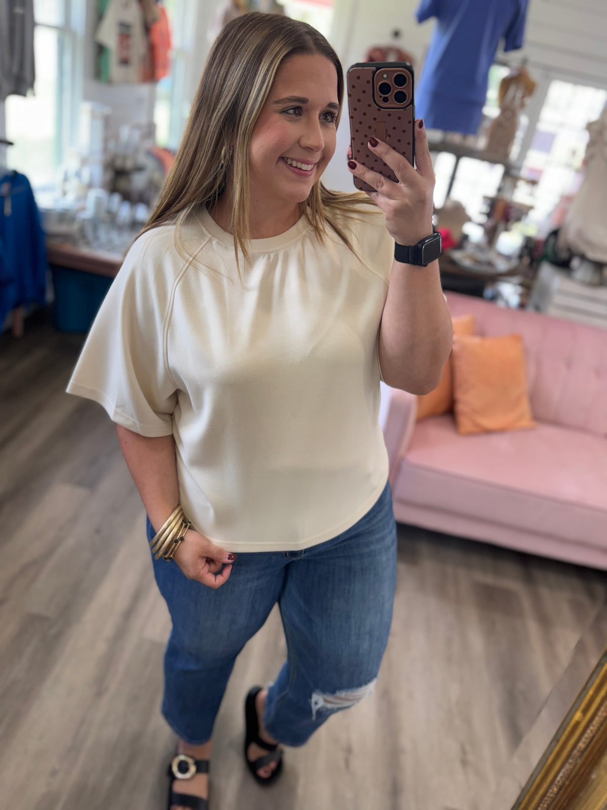 A woman takes a mirror selfie in a boutique while wearing a cream-colored short-sleeve top, blue jeans, and black sandals. The background shows a pink sofa and clothing displays.