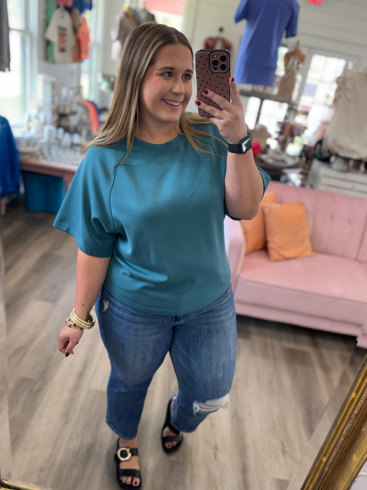 A woman takes a mirror selfie in a boutique while wearing a teal short-sleeve top and distressed blue jeans.