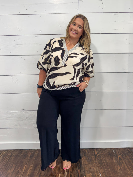 A woman modeling a short-sleeve V-neck blouse with a black and cream abstract pattern, paired with black wide-leg pants, standing against a white shiplap wall.