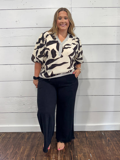 A woman modeling a short-sleeve collared blouse with a black and cream abstract leaf pattern, paired with black wide-leg pants, standing against a white shiplap wall.