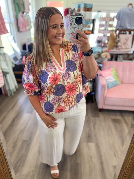 Woman taking a mirror selfie wearing a colorful floral blouse and white pants in a store.